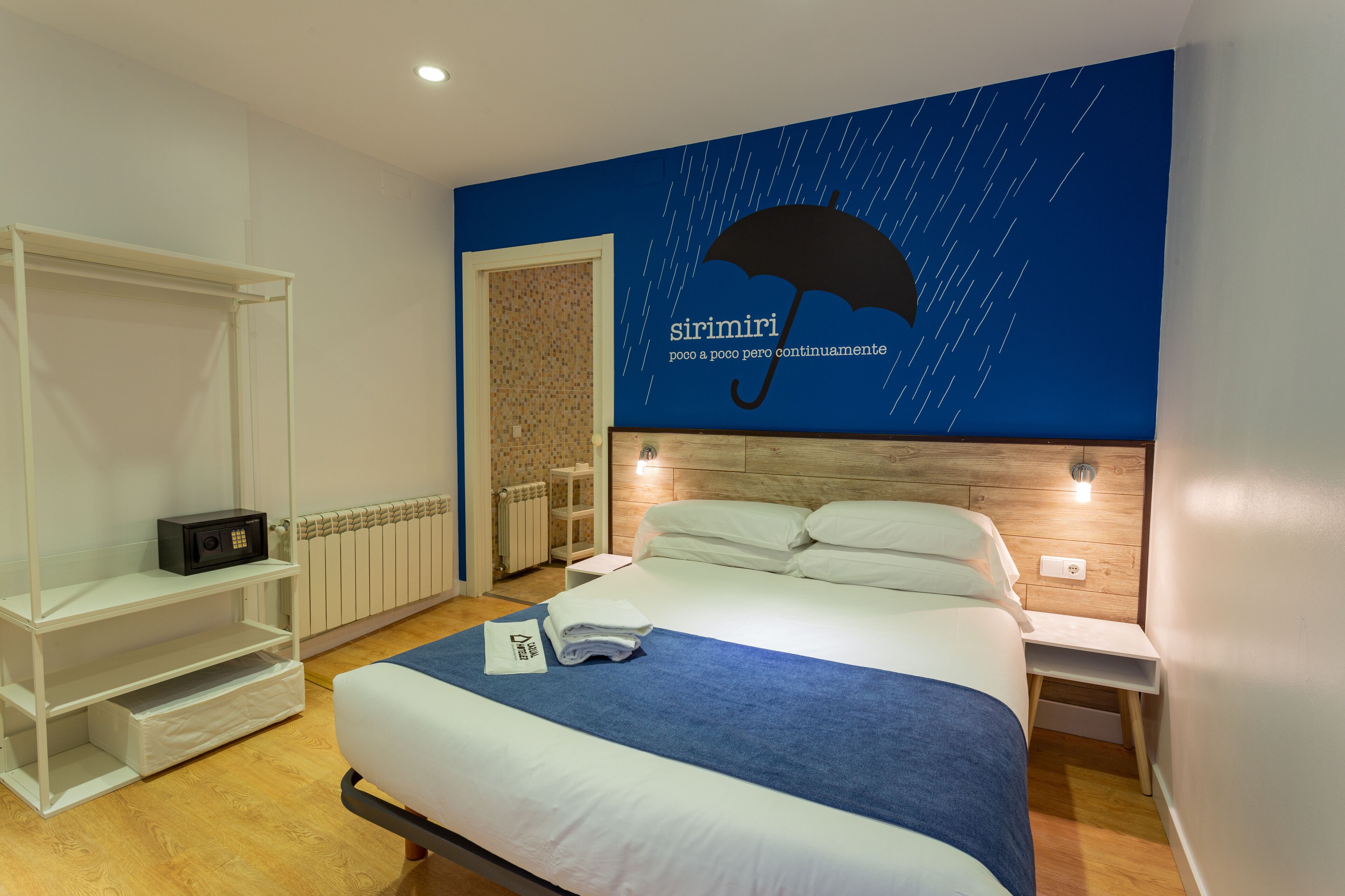 A hotel room with a bed, a nightstand, a shelf, and a wall with a rain umbrella design.