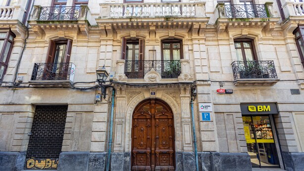 A building with a large wooden double door, a BM store on the right, and a small balcony with a plant. - Casual Blue (Bilbao)
