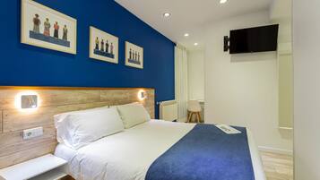 Una habitación de hotel moderna con cabecera de madera, una cama grande con ropa de cama blanca y azul, una mesita de noche, un televisor de pantalla plana montado en la pared y una obra enmarcada sobre la cama.