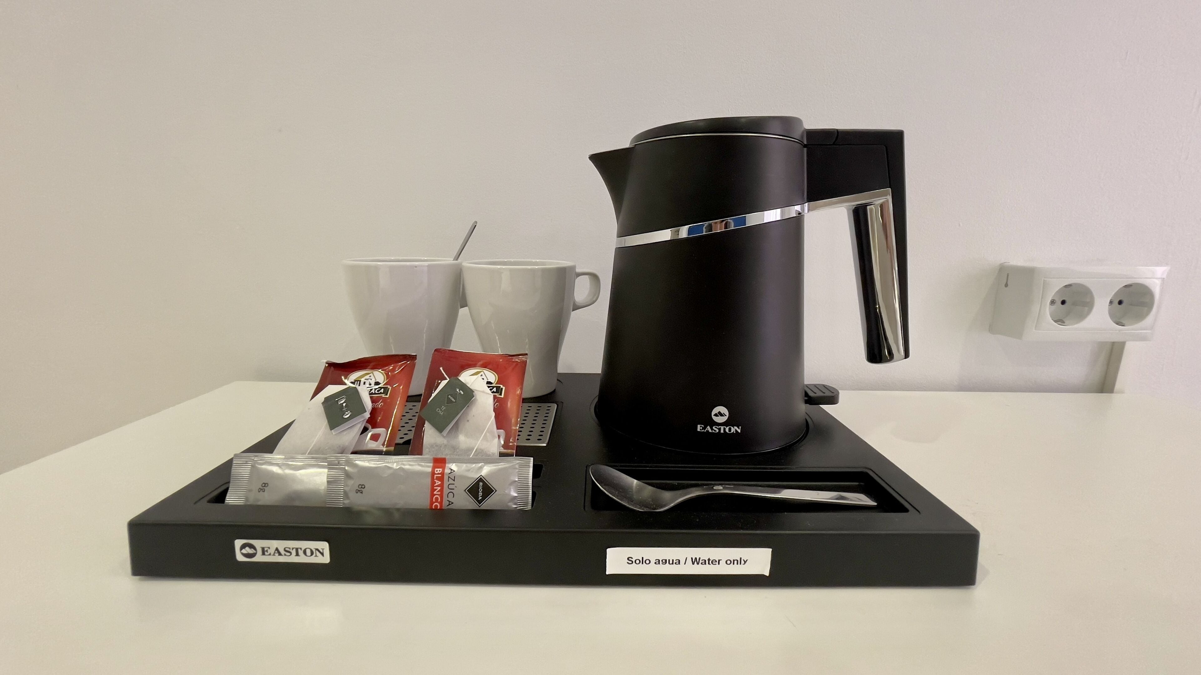 A black coffee maker with a water reservoir, two white cups, and several packets of coffee and creamer on a tray.