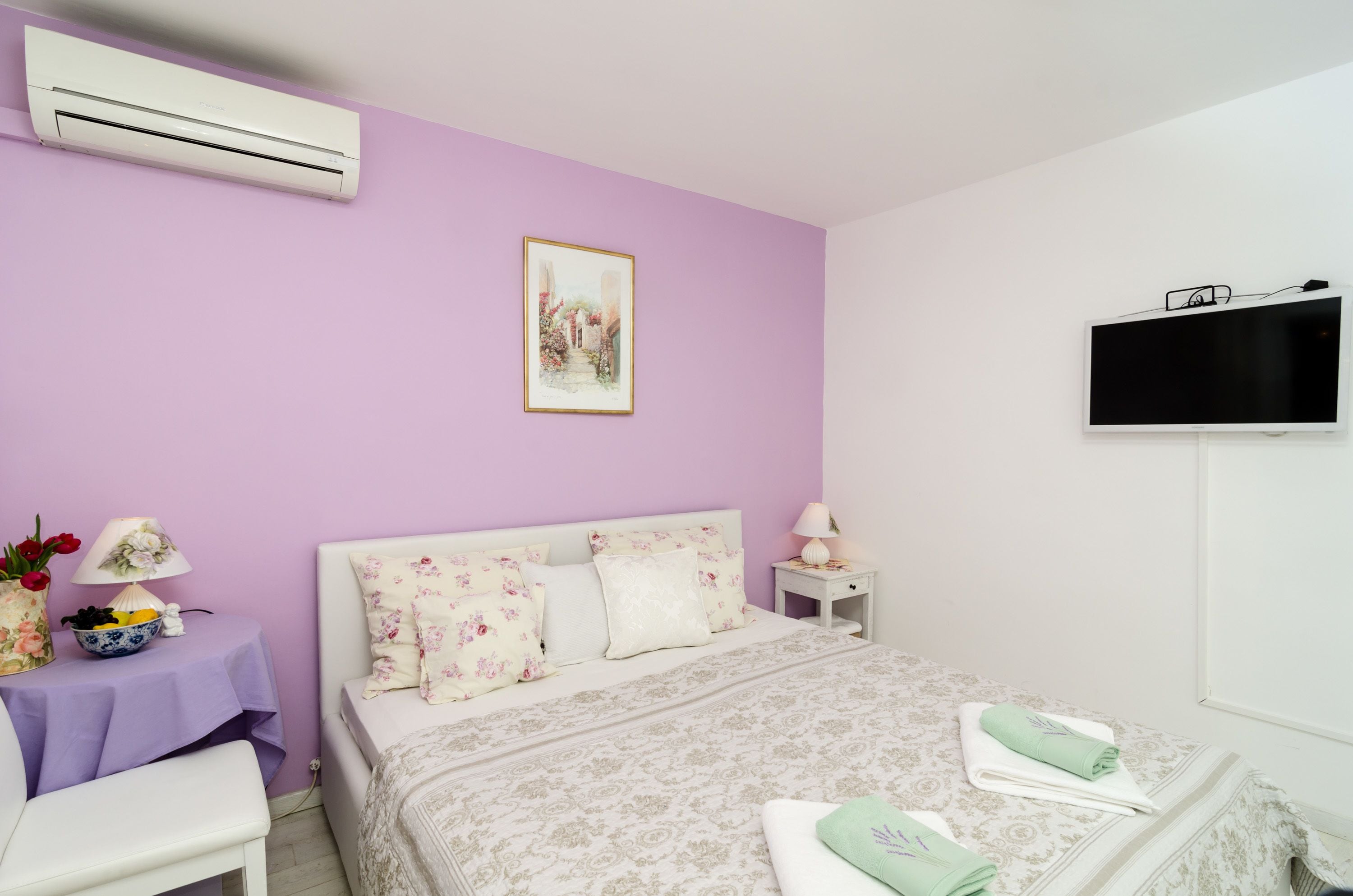 A bedroom with a bed, a bedside table with a lamp, a small table with a vase of flowers, and an air conditioning unit on the wall.