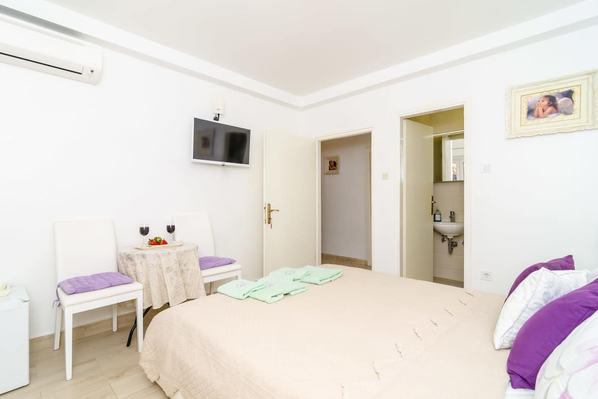 Quarto com cama, cadeira, mesa pequena com bandeja de copos de vinho e televisão na parede.