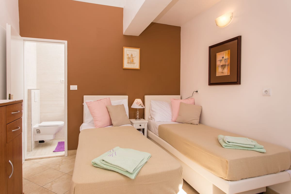 Quarto de hotel com duas camas, banheiro e uma pequena mesa com luminária.