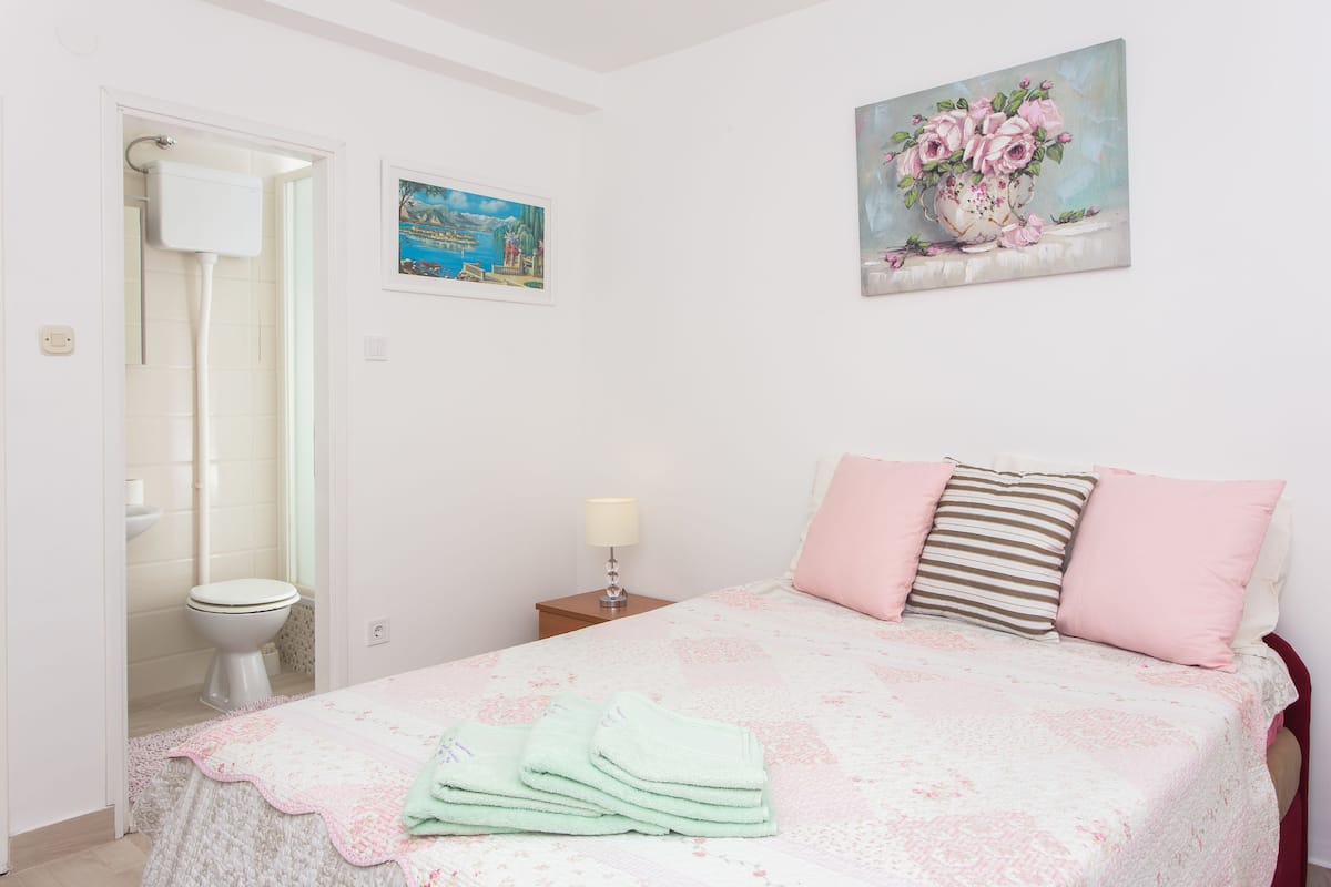Quarto com cama, almofadas, mesa de cabeceira, pintura e banheiro com vaso sanitário e chuveiro.
