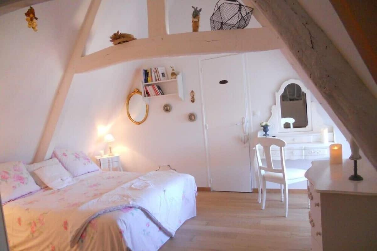 Un dormitorio con cama, mesita de noche, lámpara y un escritorio con silla. Hay una estantería y un espejo en la pared.