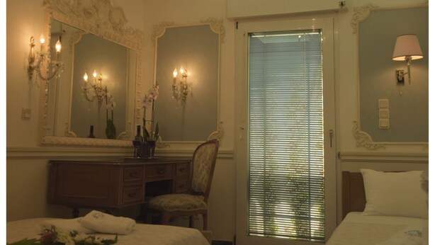 A hotel room with a large bed, a smaller bed, a vanity with a mirror, a chandelier, and a window with blinds. - Hotel Esperides (Naxos)