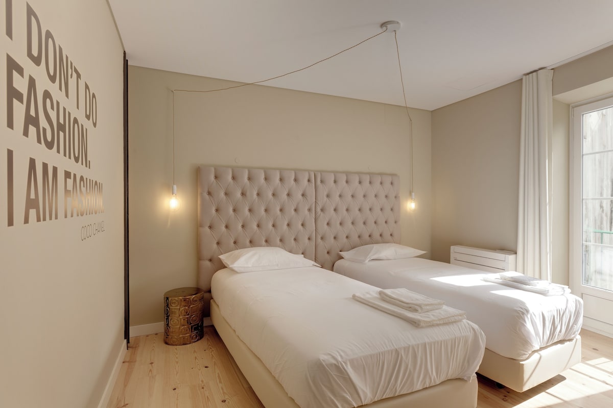 A modern bedroom with two beds, a tufted headboard, a wall-mounted light, and a window with curtains.