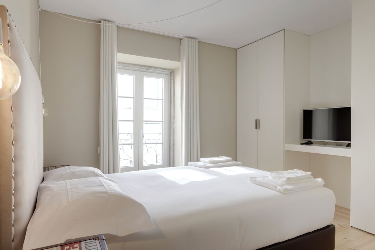 A modern hotel room with a large bed, a TV on a stand, and a window with curtains.