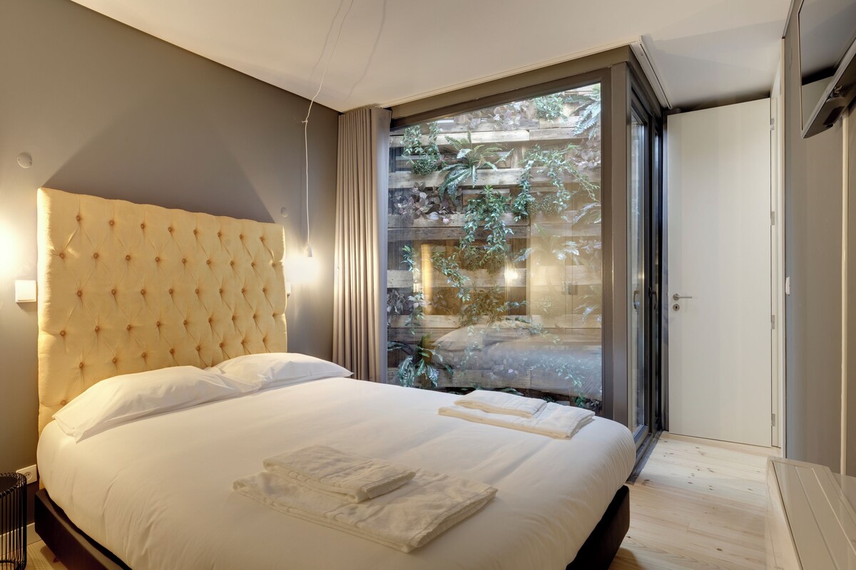 A bedroom with a large bed, a wooden headboard, a sliding glass door leading to a balcony with plants, and a white wall.