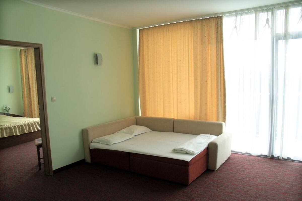 A hotel room with a single bed, a small stool, a mirror, and a window with curtains.