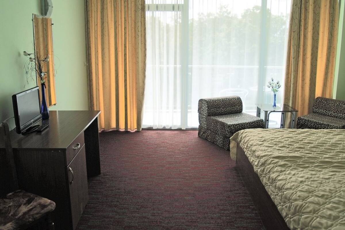 A hotel room with a bed, two armchairs, a desk with a television, and a large window with curtains.