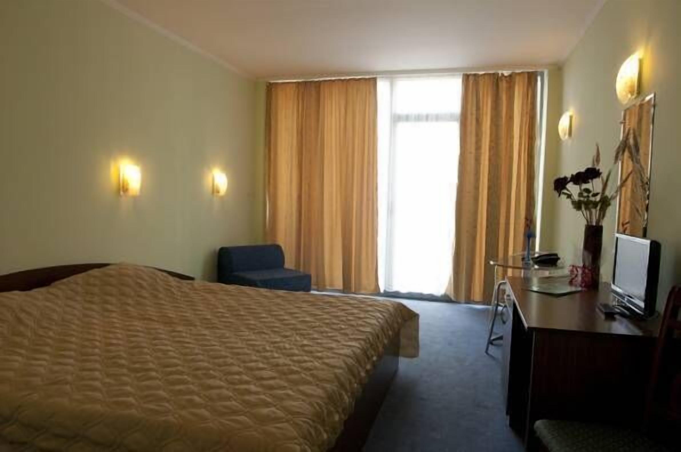 A hotel room with a large bed, a desk with a computer, a chair, and a window with curtains.
