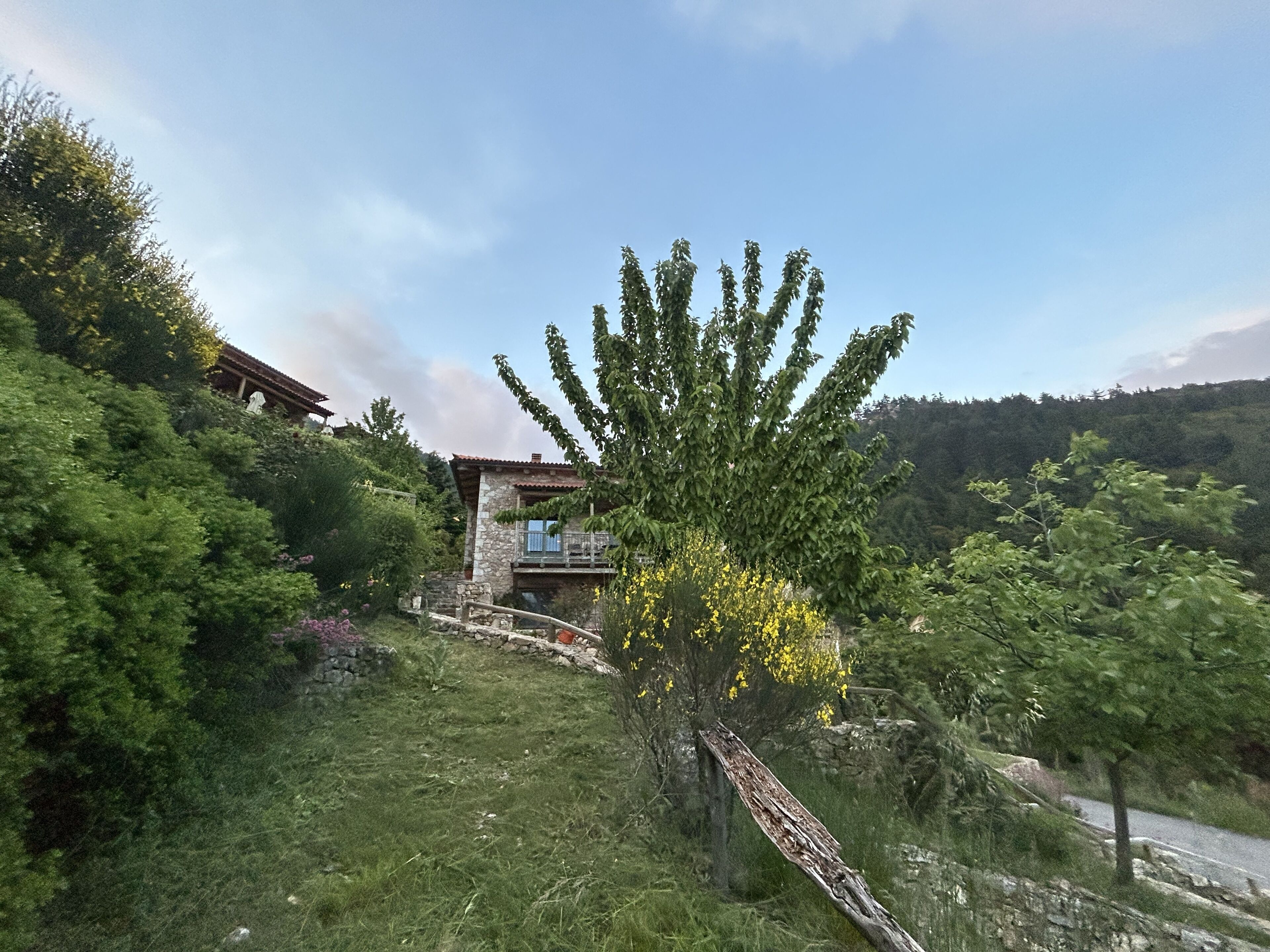 A stone house with a balcony, surrounded by lush greenery and a forested hillside.