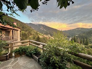 A stone building with a balcony, surrounded by lush greenery and mountains in the background. - Pritanio (Sparta)
