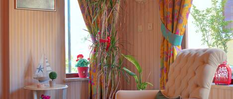 A room with striped wallpaper, a white armchair, a small table with a plant, and a window with colorful curtains.