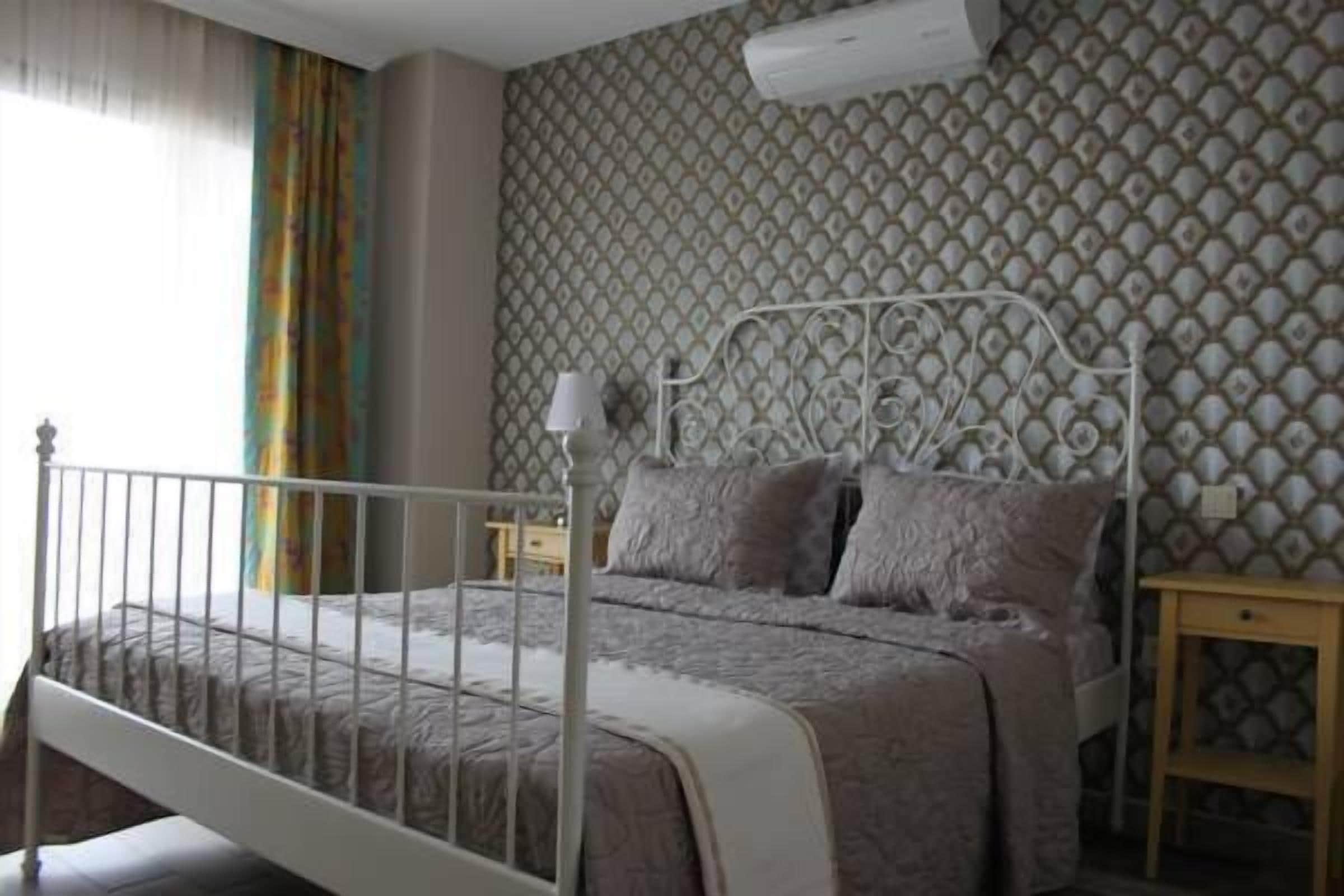 A bedroom with a white metal bed frame, a patterned wallpaper, a bedside table, and a nightstand.