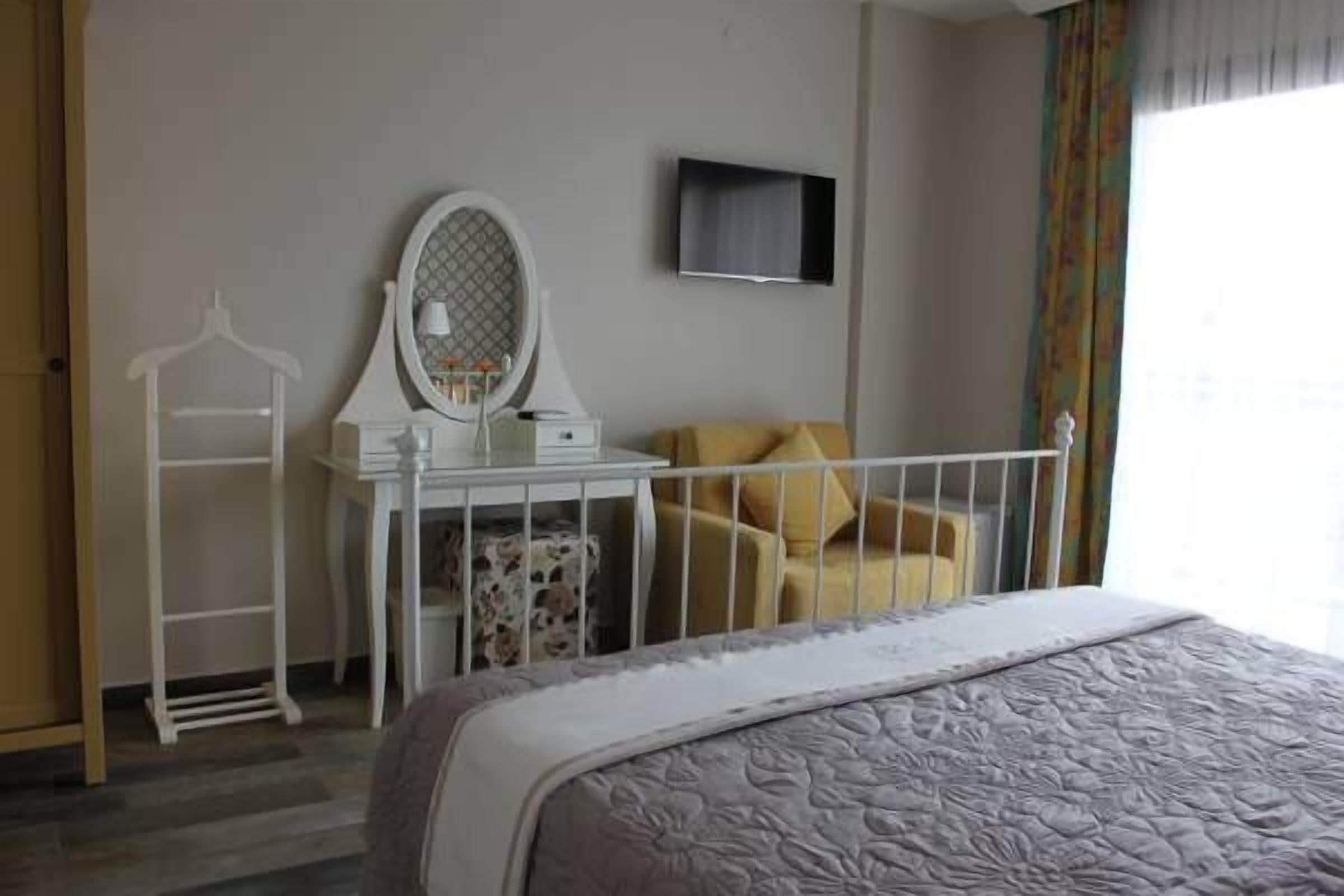 A hotel room with a bed, a white dresser with a mirror, a television on the wall, and a window with curtains.
