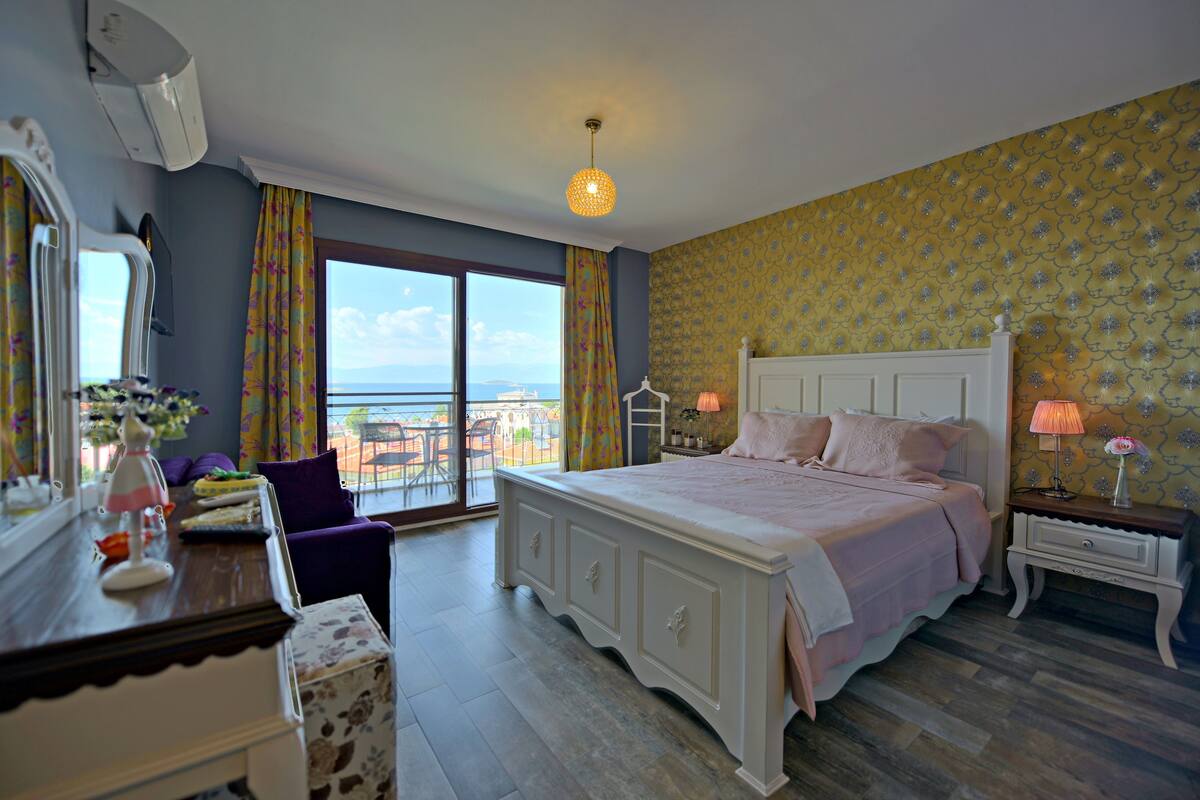 A bedroom with a large bed, a dresser, a vanity, and a view of the sea.
