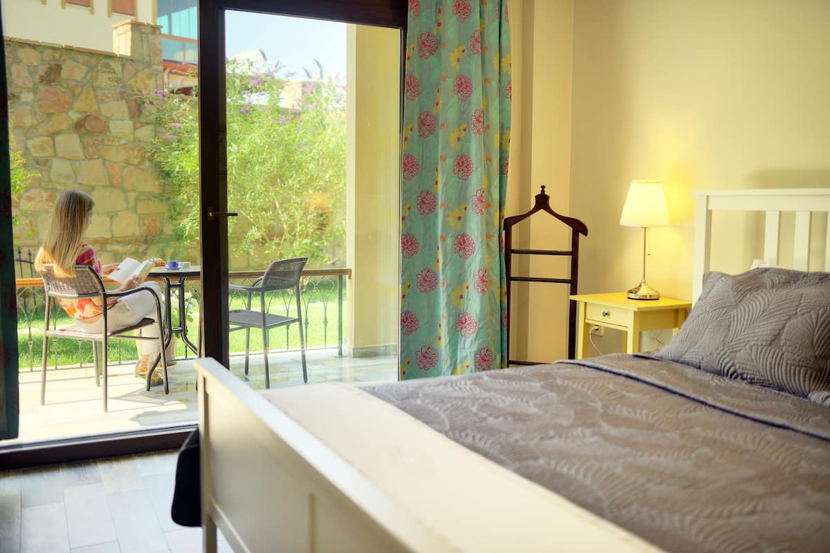 A bedroom with a bed, a bedside table with a lamp, a chair, and a view of a balcony with a person reading a book.