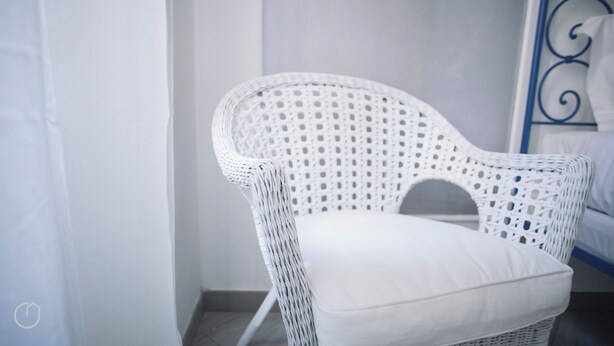 A white wicker chair with a cushion in a room with a white wall and a metal bed frame. - Italianway   - C.so Sempione 96 (Milan)
