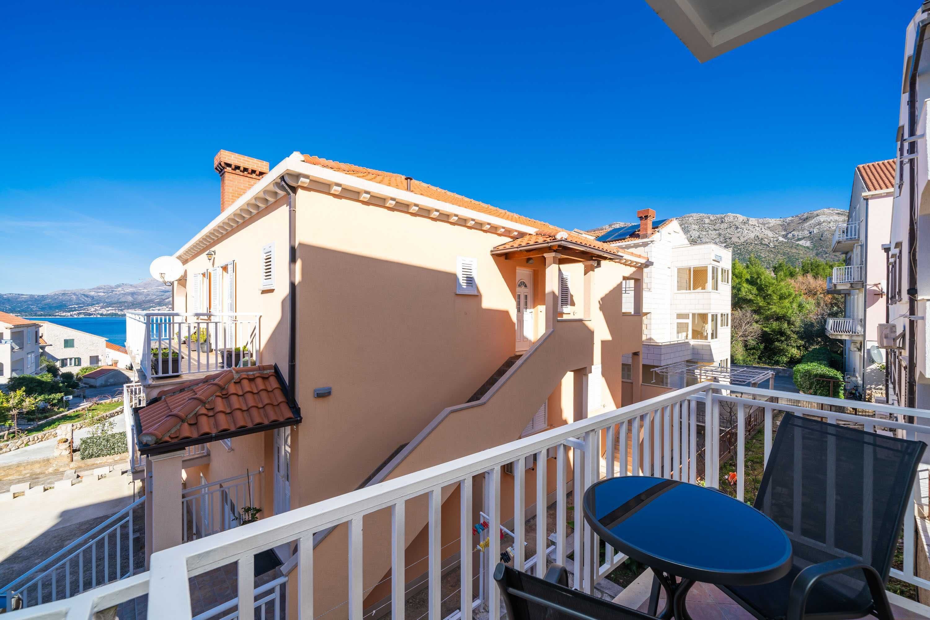 A balcony with a table and chairs, overlooking a coastal area with buildings and a mountain in the distance.
