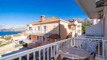Un balcone con tavolo e sedie, affacciato su una zona costiera con edifici e un cielo azzurro limpido.