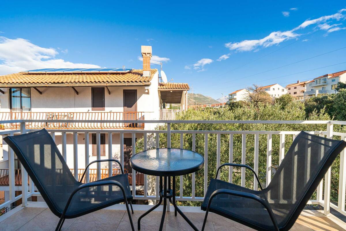 A balcony with a round table and two chairs, overlooking a residential area with terracotta-tiled roofs and greenery.