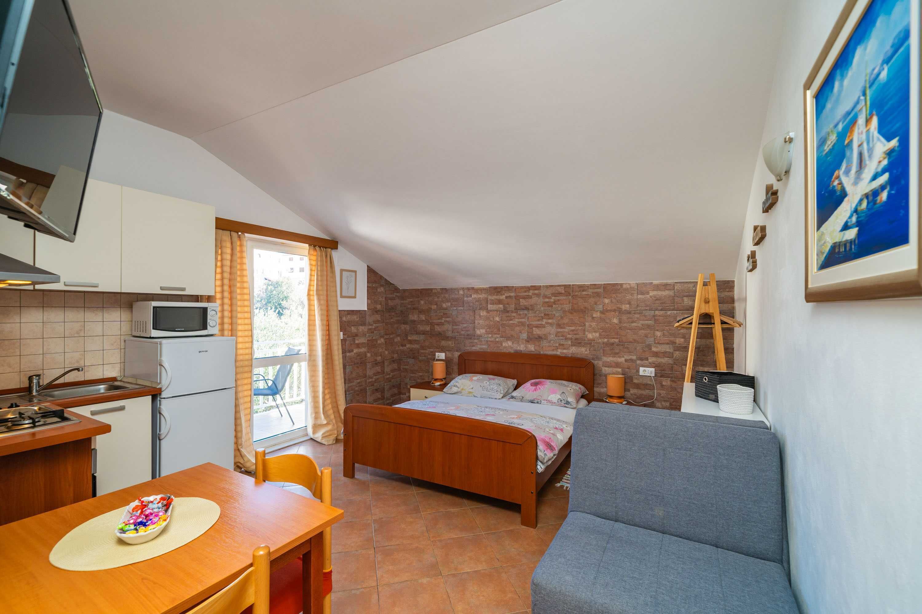 A compact room with a kitchenette, a bed, a sofa, and a window with curtains.