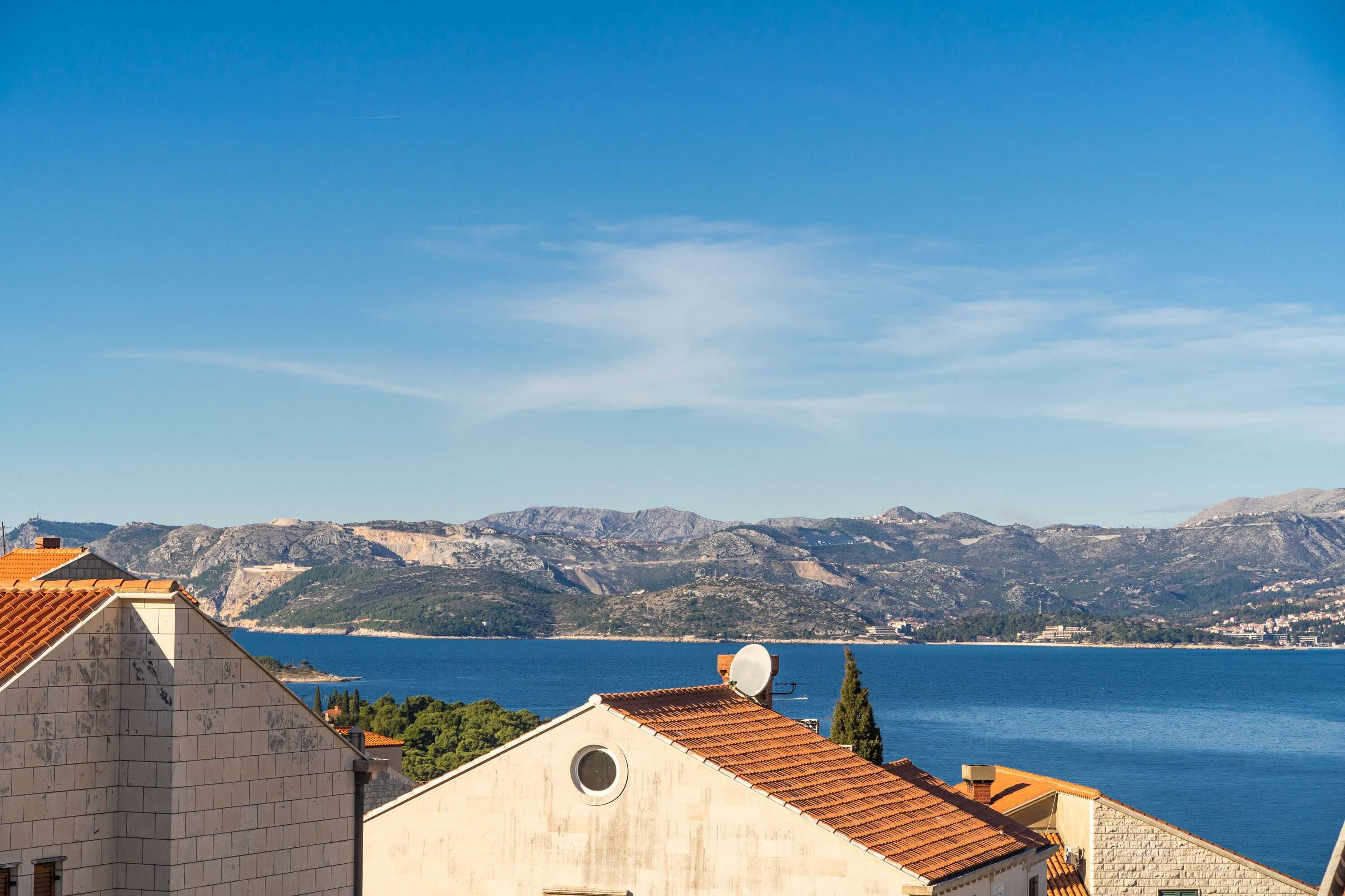 A coastal town with terracotta-roofed buildings and a clear view of the sea and mountains in the background.