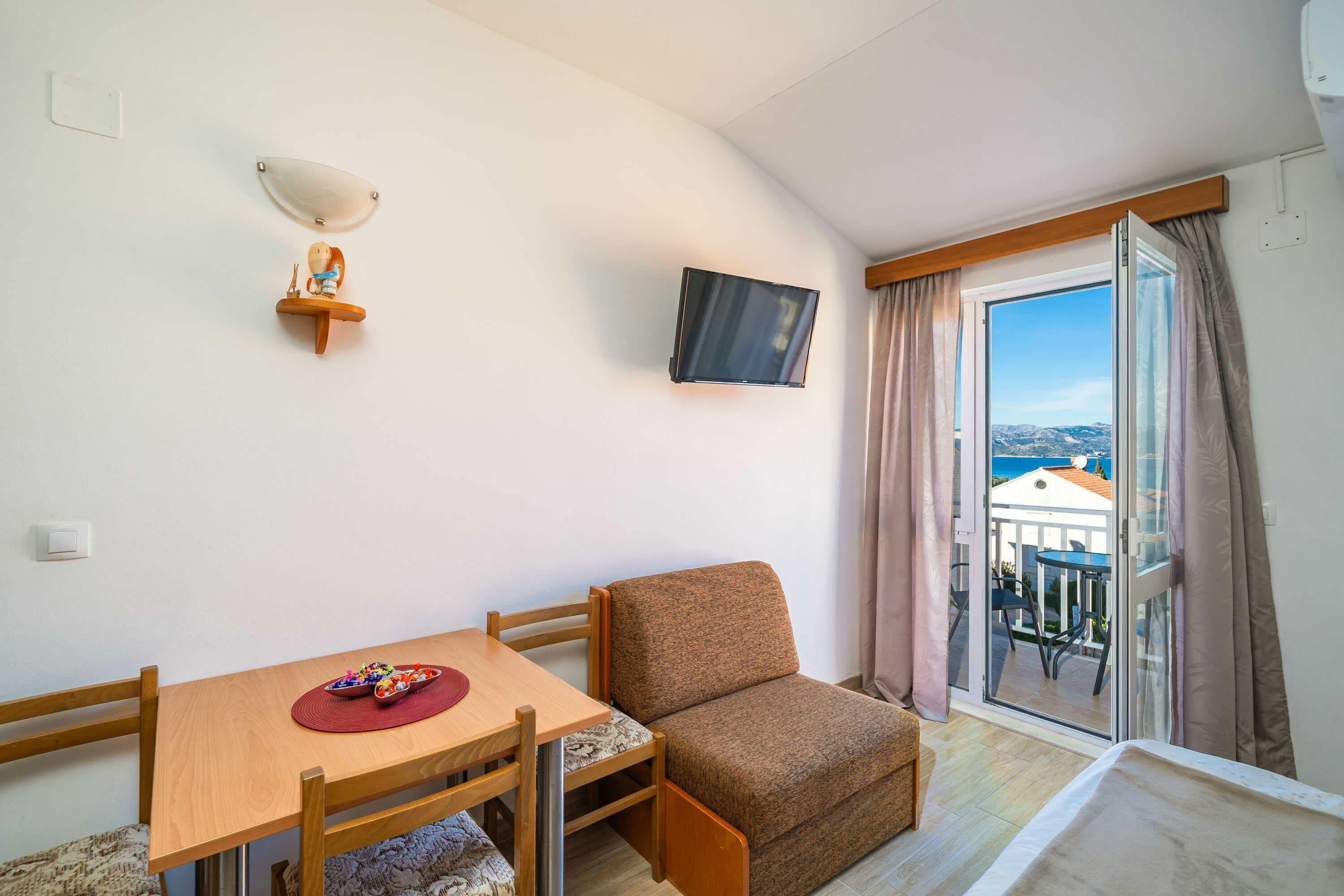 Ein kleines Zimmer mit einem Holztisch, einer Couch, einem an der Wand montierten Fernseher und einer Glastür zum Balkon.