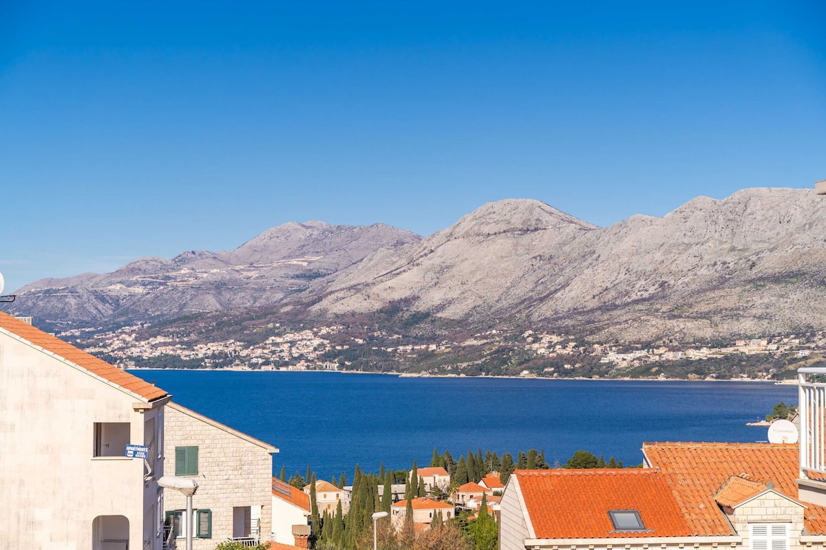 A coastal town with buildings featuring red-tiled roofs and a clear view of the sea and mountains in the background.