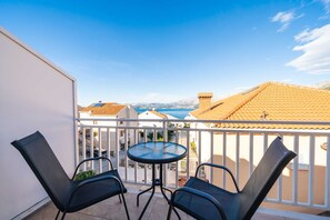 Un balcon avec deux chaises et une petite table, donnant sur une zone côtière avec des bâtiments et des montagnes à l’horizon.