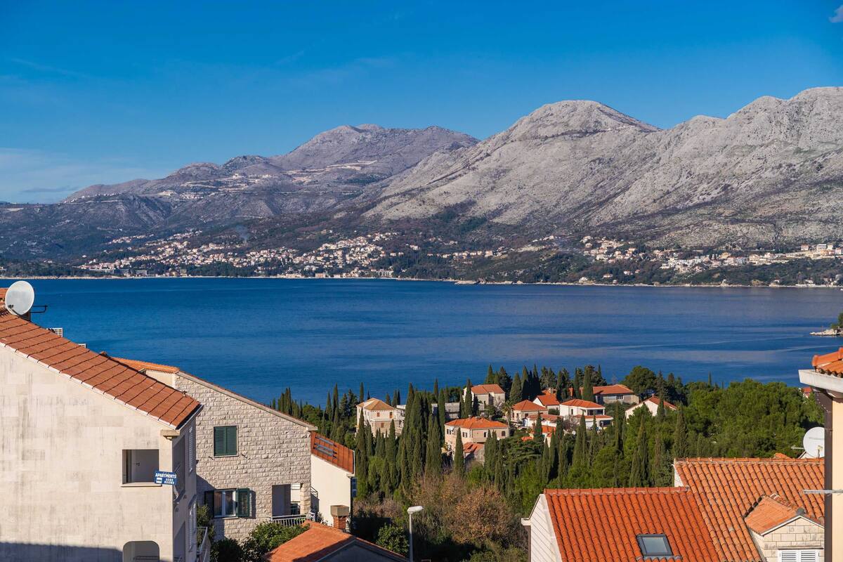 A coastal town with stone buildings, a clear blue sea, and mountains in the background.