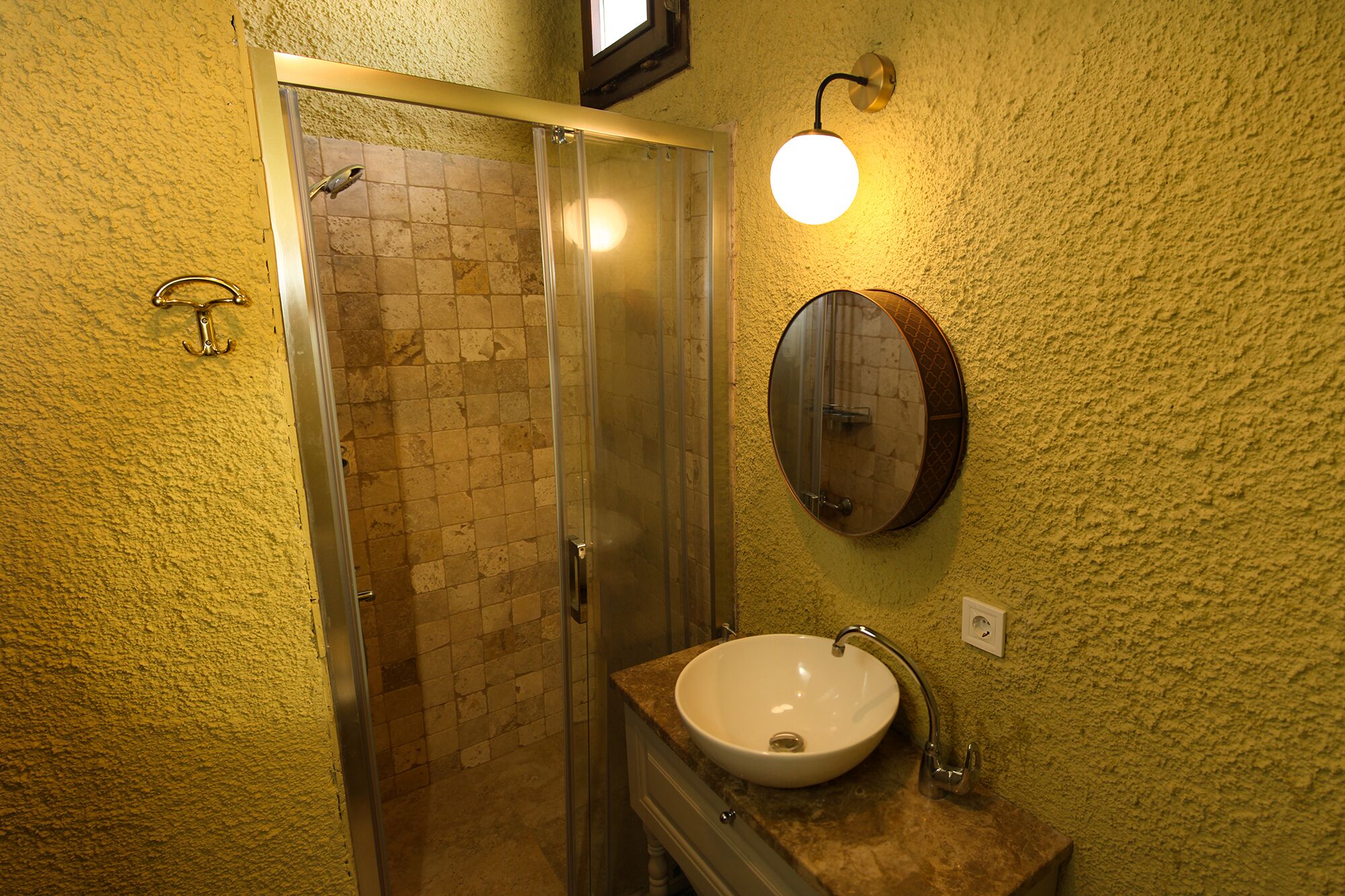 Ein Badezimmer mit einer Duschkabine aus Glas, einem weißen Waschbecken und einem runden Spiegel an einer strukturierten Wand.