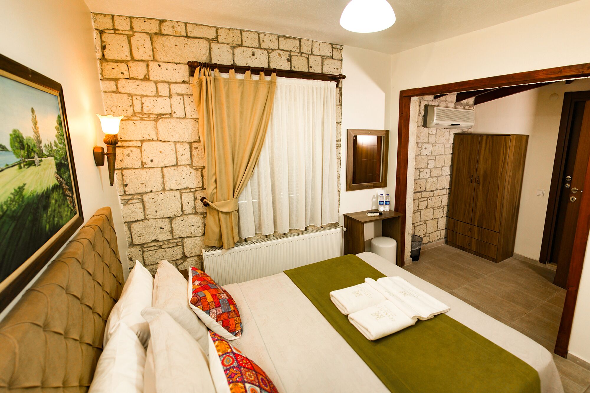 A hotel room with a brick wall, a bed with white bedding and a green throw, a window with curtains, a bedside table with a lamp, a mirror, and a wooden wardrobe.