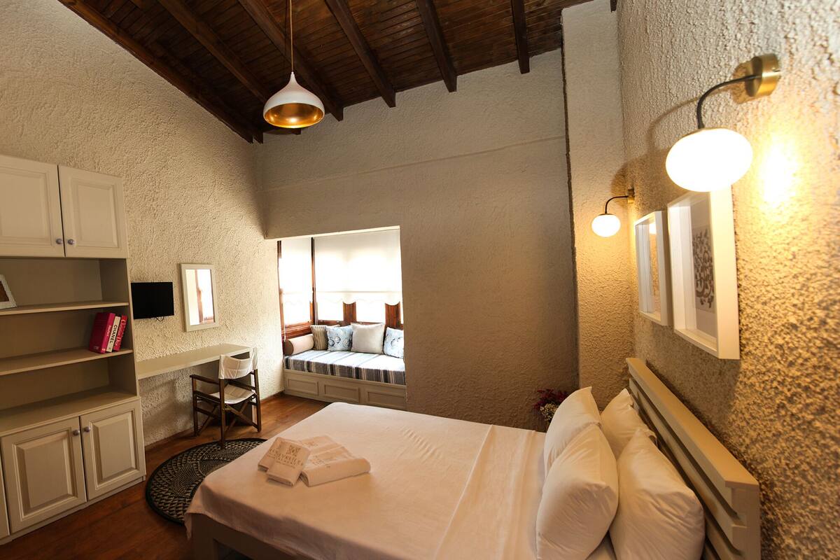 Un dormitorio con cama, escritorio y silla. Hay libros en el escritorio y una ventana con vistas al exterior.
