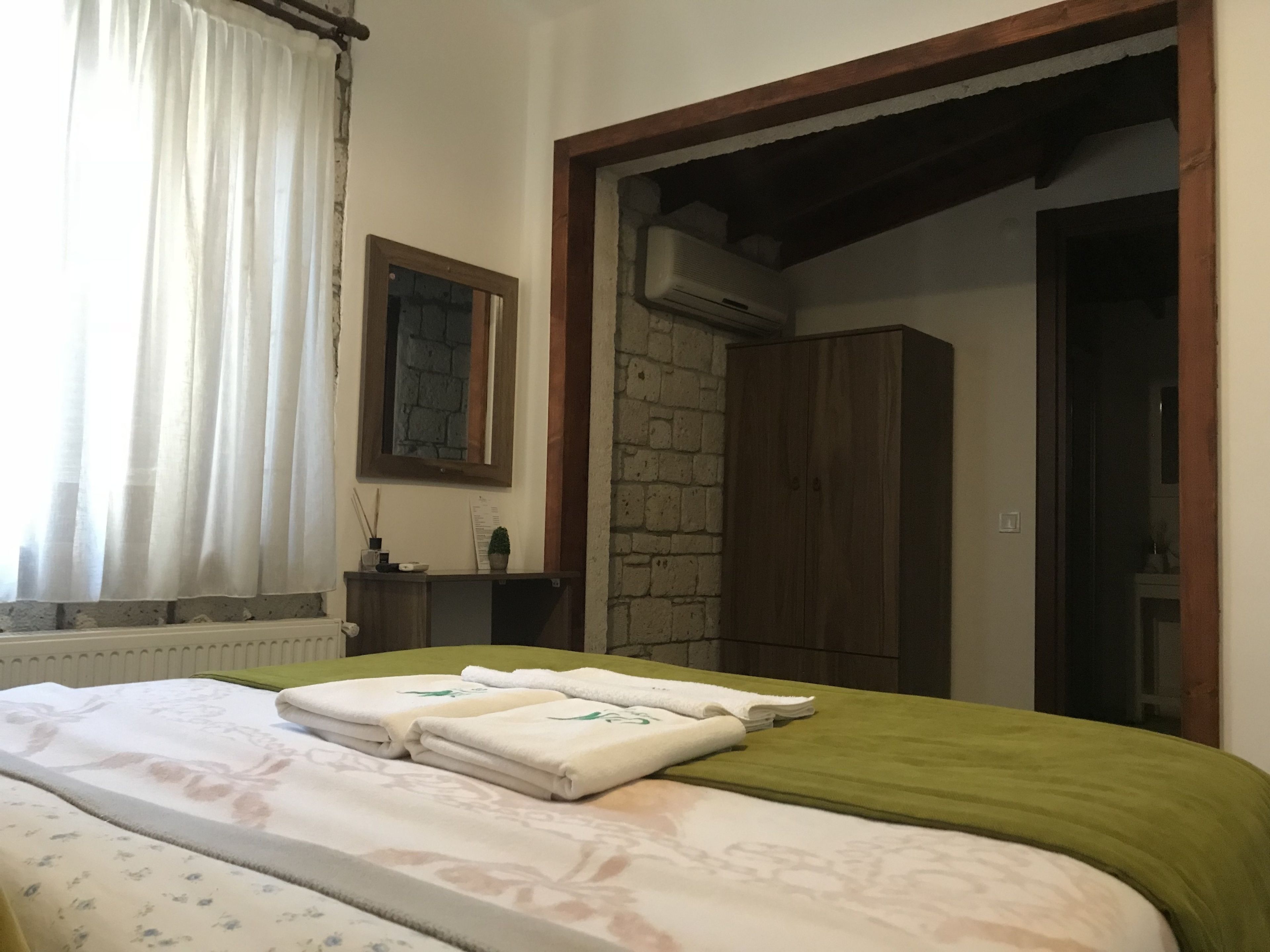 A hotel room with a bed, a green bedspread, a wooden wardrobe, a stone wall, and a window with white curtains.