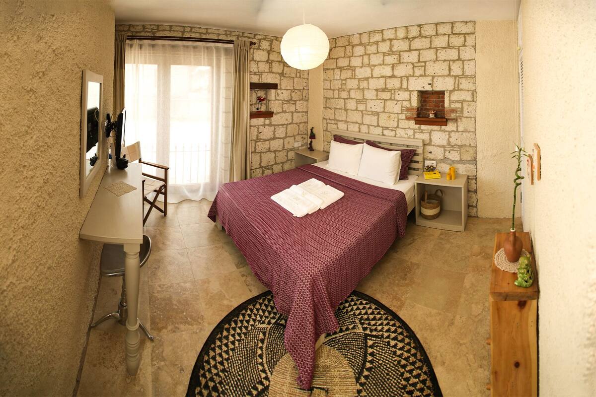 Un dormitorio con pared de ladrillo, una cama con colcha de cuadros rojos y blancos, una mesita de noche con lámpara y un pequeño estante con objetos decorativos.
