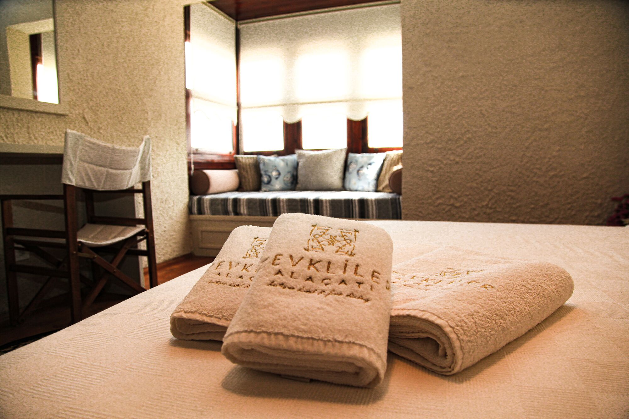A neatly made bed with folded towels and a chair in a room with a window and a sofa.