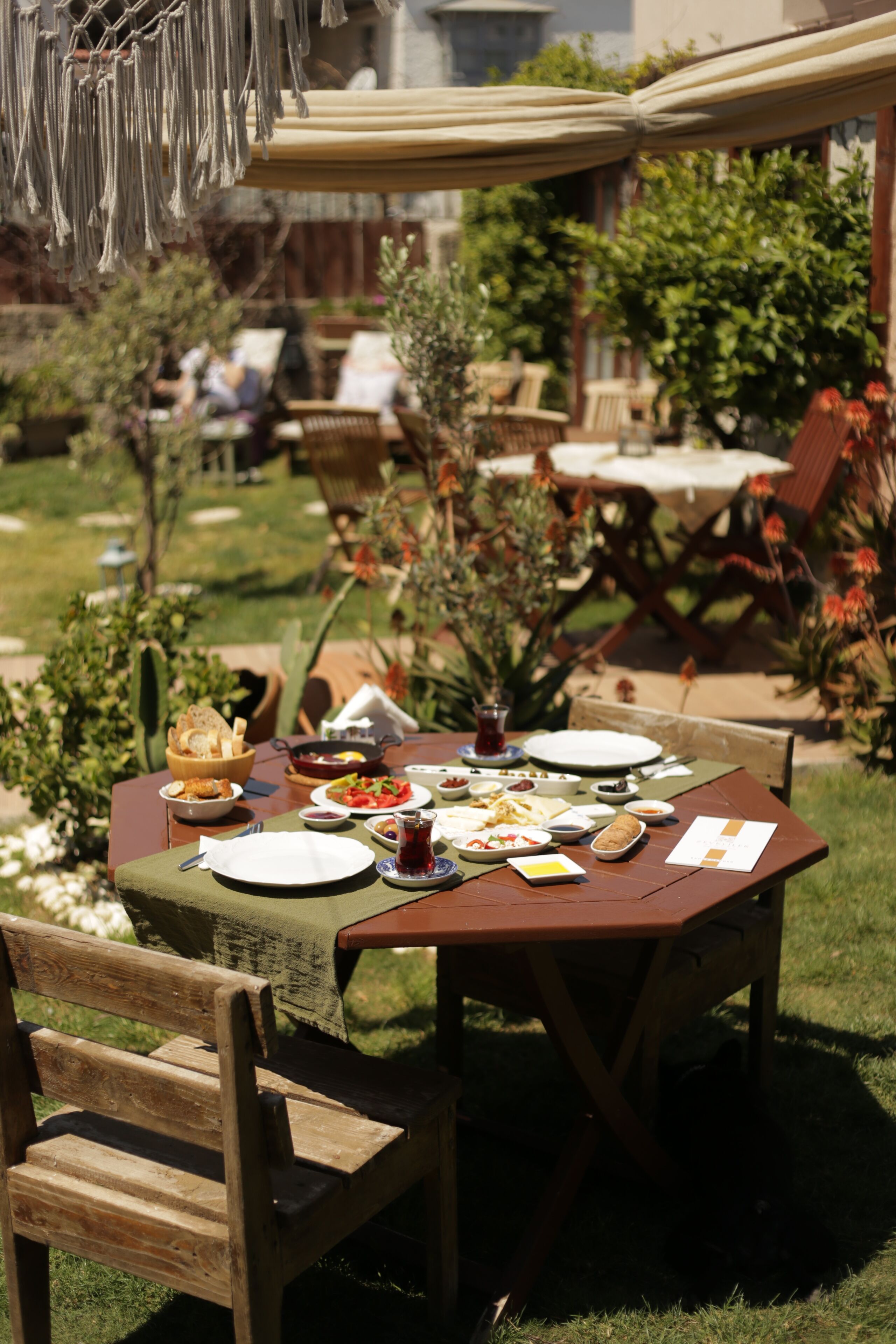 Una mesa de madera al aire libre con varios platos y una bebida, rodeada de sillas y follaje.