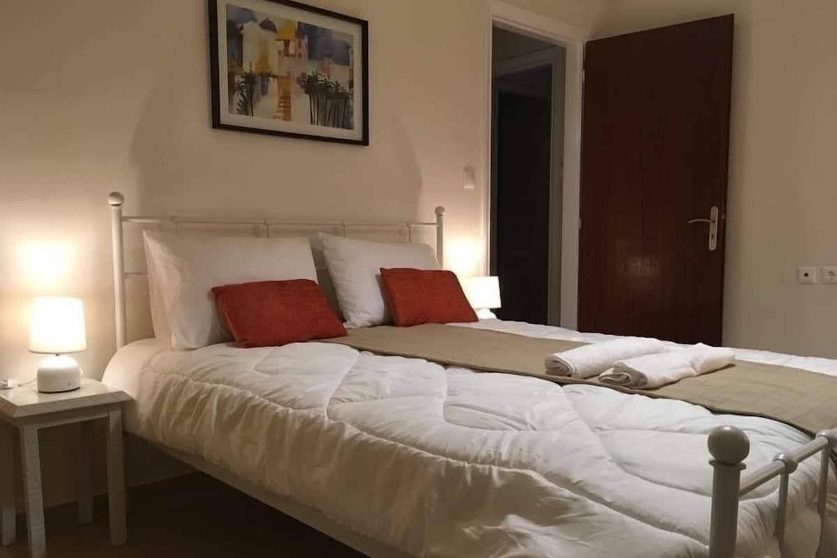 A neatly made bed with white bedding and red pillows, a bedside lamp, and a framed picture on the wall.