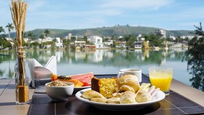 Una mesa de desayuno con diversos alimentos, un vaso de jugo de naranja y una vista de un lago con edificios al fondo.