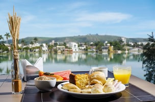 Mesa de café da manhã com diversos alimentos, um copo de suco de laranja e vista para um lago com edifícios ao fundo.