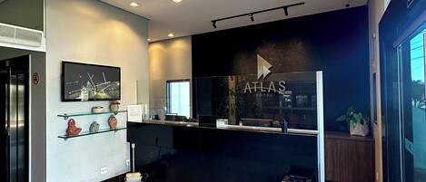 Uma área de recepção de hotel moderna com um balcão preto, uma televisão e uma placa com a inscrição 'ATLAS HOTEL'.