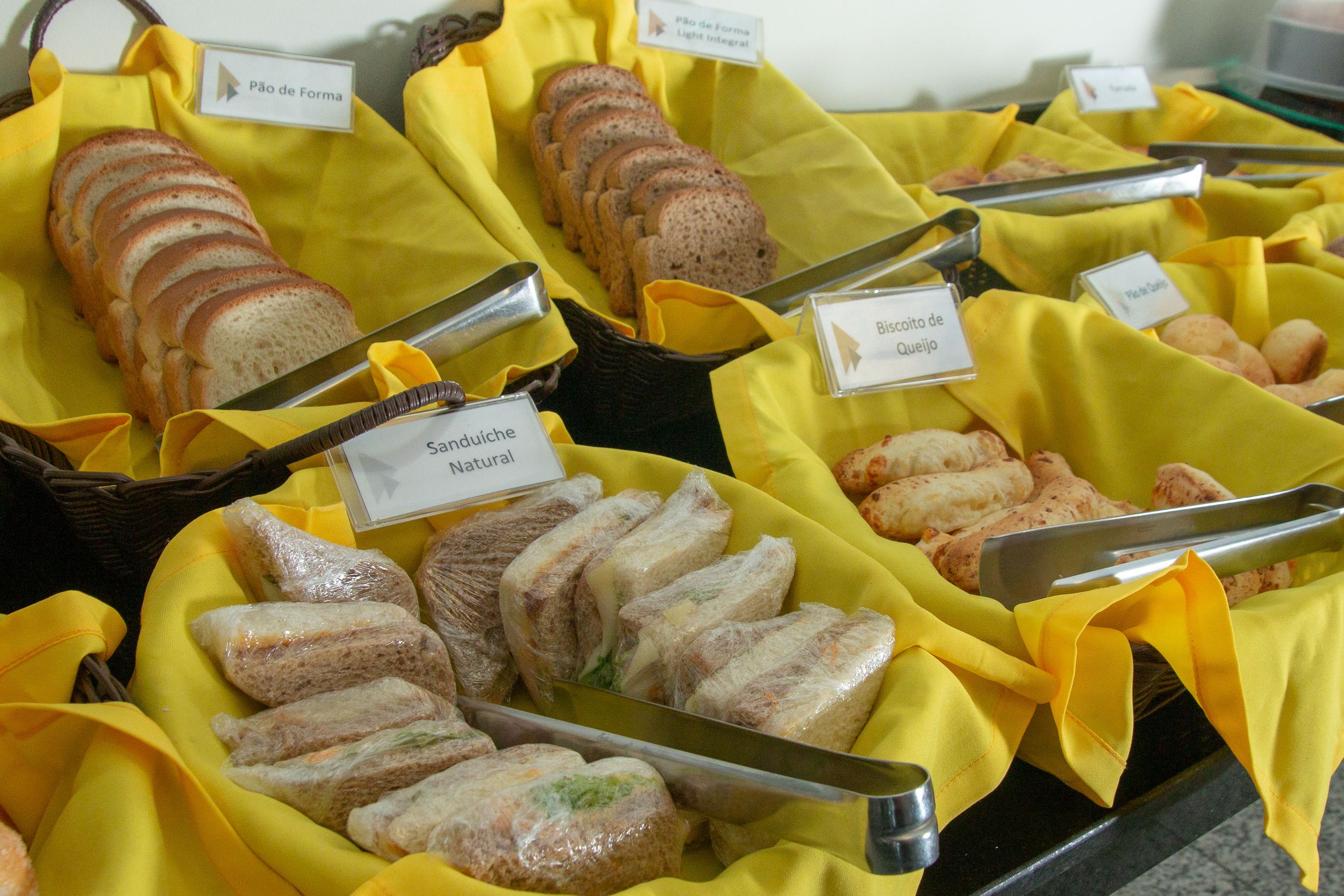 Tarjolla on monipuolinen buffet, jossa on erilaisia ruokia, kuten voileipiä ja leipää. Ruokalajit on merkitty nimilapuilla, esimerkiksi ”Sanduiche Natural” ja ”Biscoito de Queijo”.