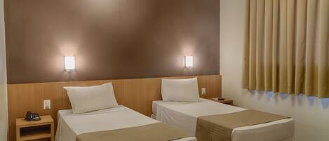A hotel room with two single beds, a wooden headboard, bedside lamps, a nightstand with a phone, and a window with curtains.