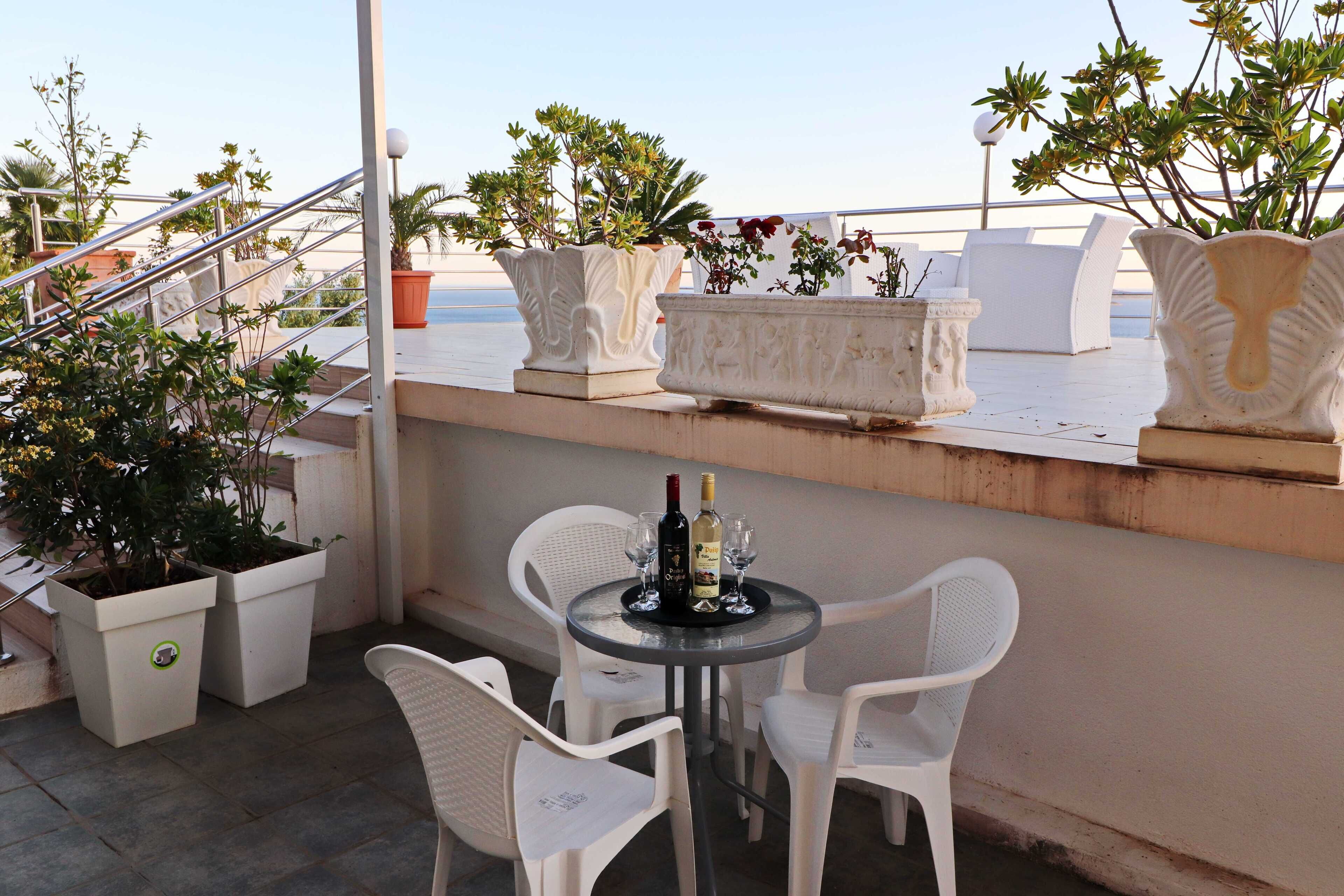 A terrace with white chairs, a table with a bottle and glasses, and potted plants.
