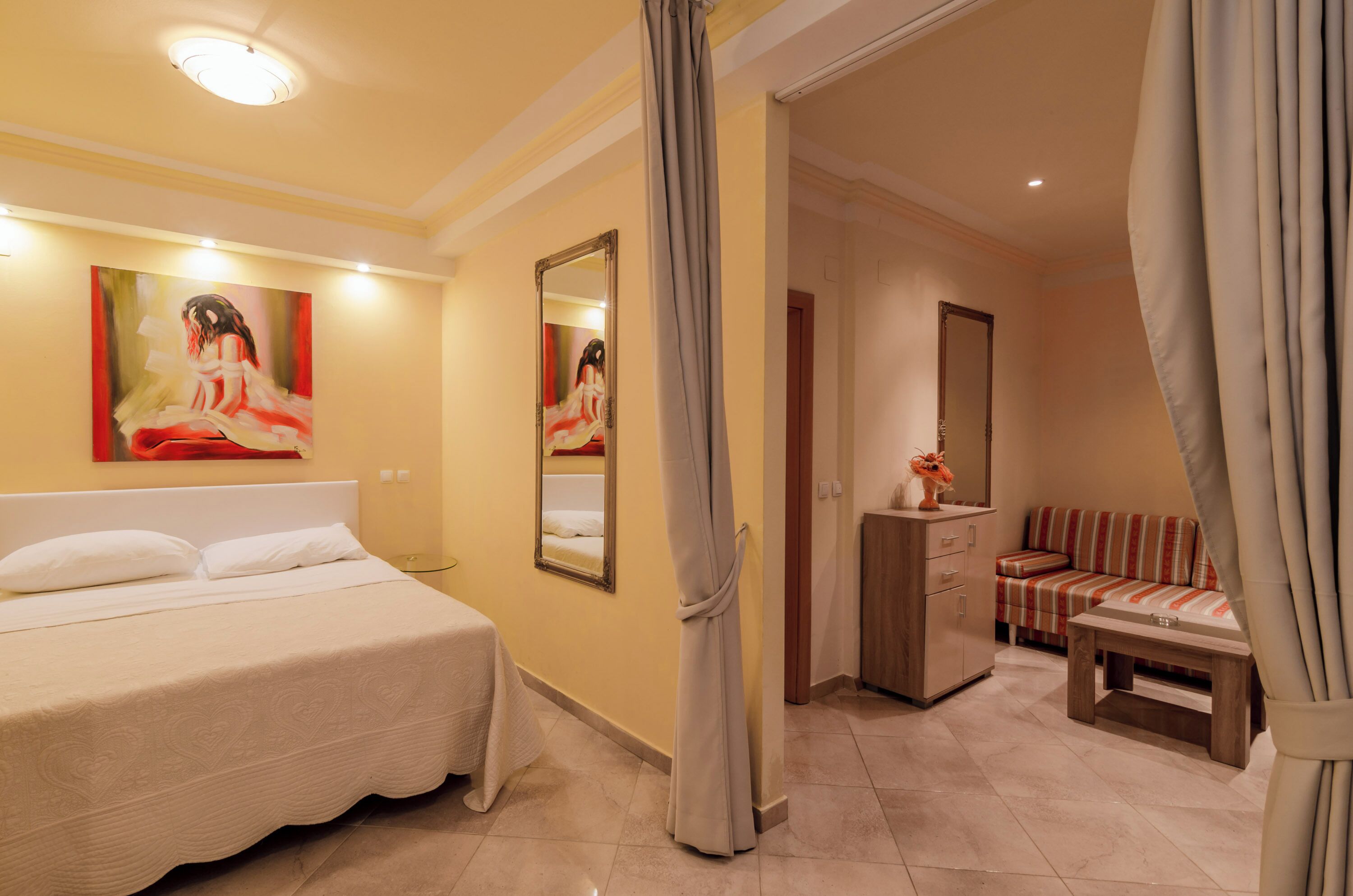 A hotel room with a bed, a sofa, a small table, a mirror, and a painting on the wall.