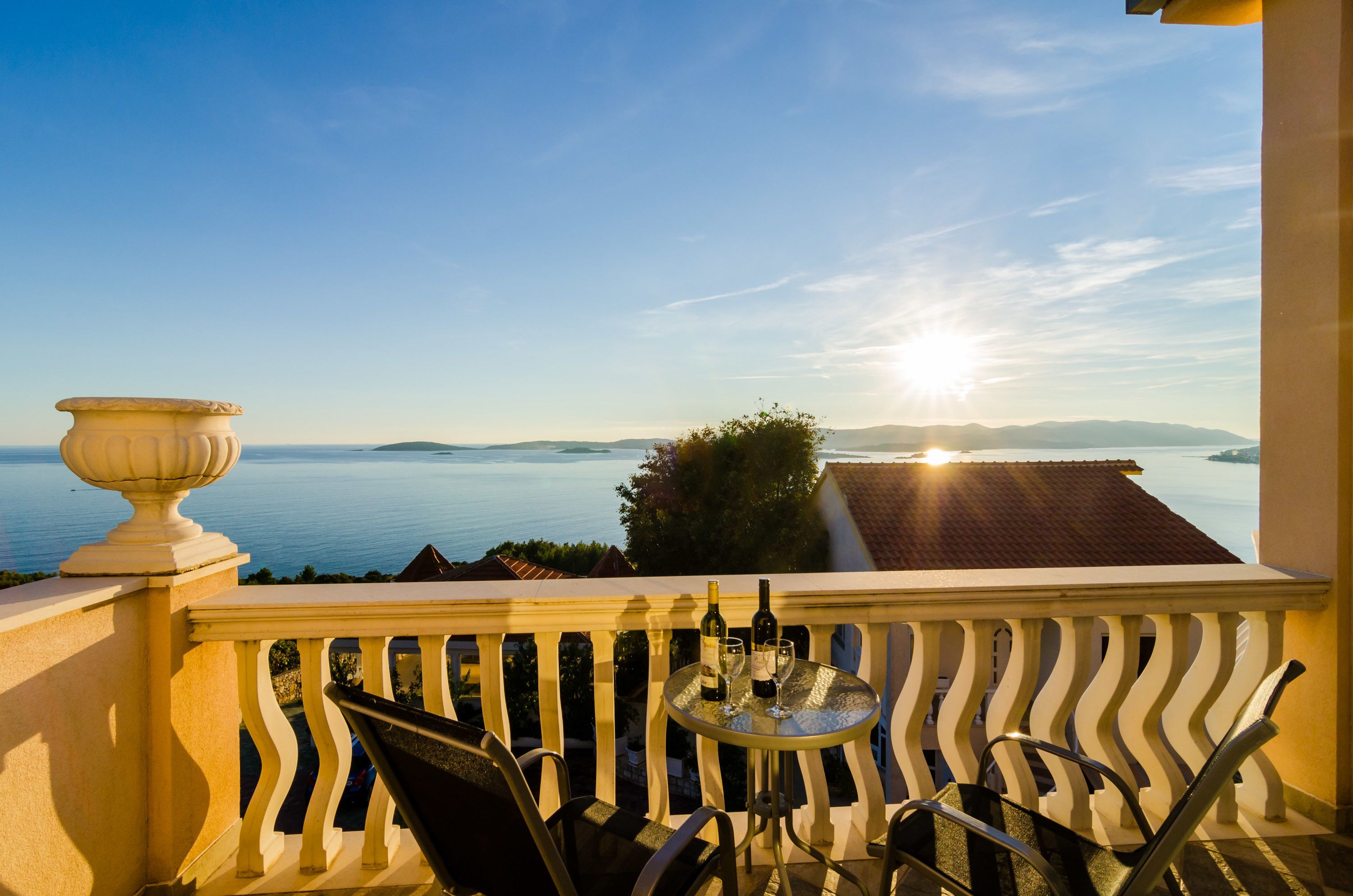 A balcony with a view of the sea, a table with glasses and a bottle, and a decorative urn.