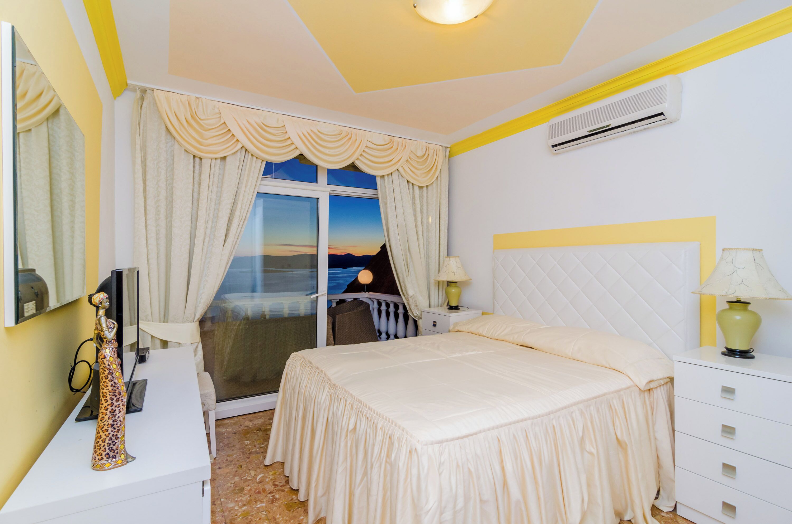 A hotel room with a bed, bedside tables, a lamp, a window with curtains, and a view of the sunset.
